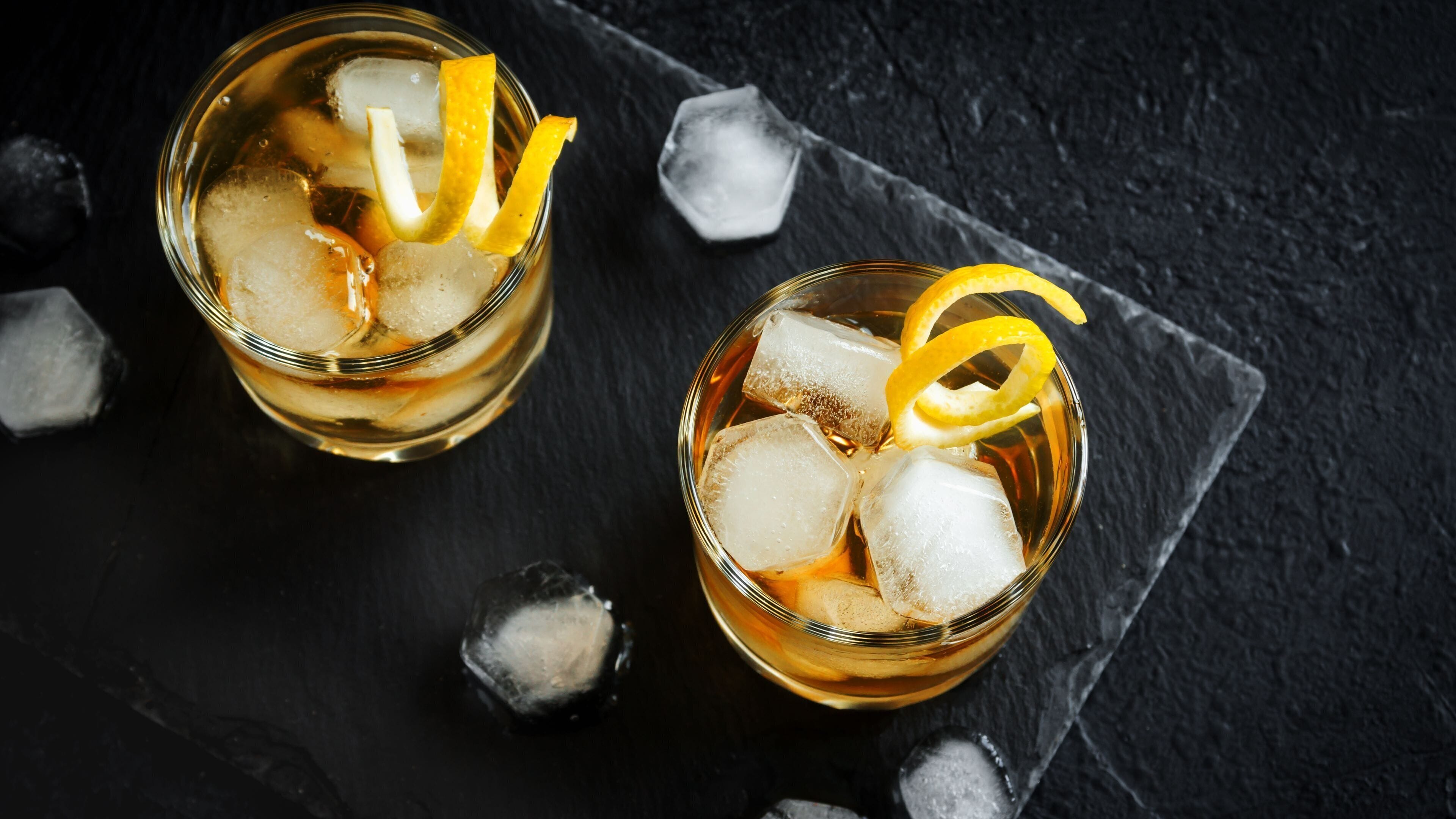 Dos vasos de líquido ámbar con cubitos de hielo y una cáscara de limón sobre una superficie de pizarra oscura.