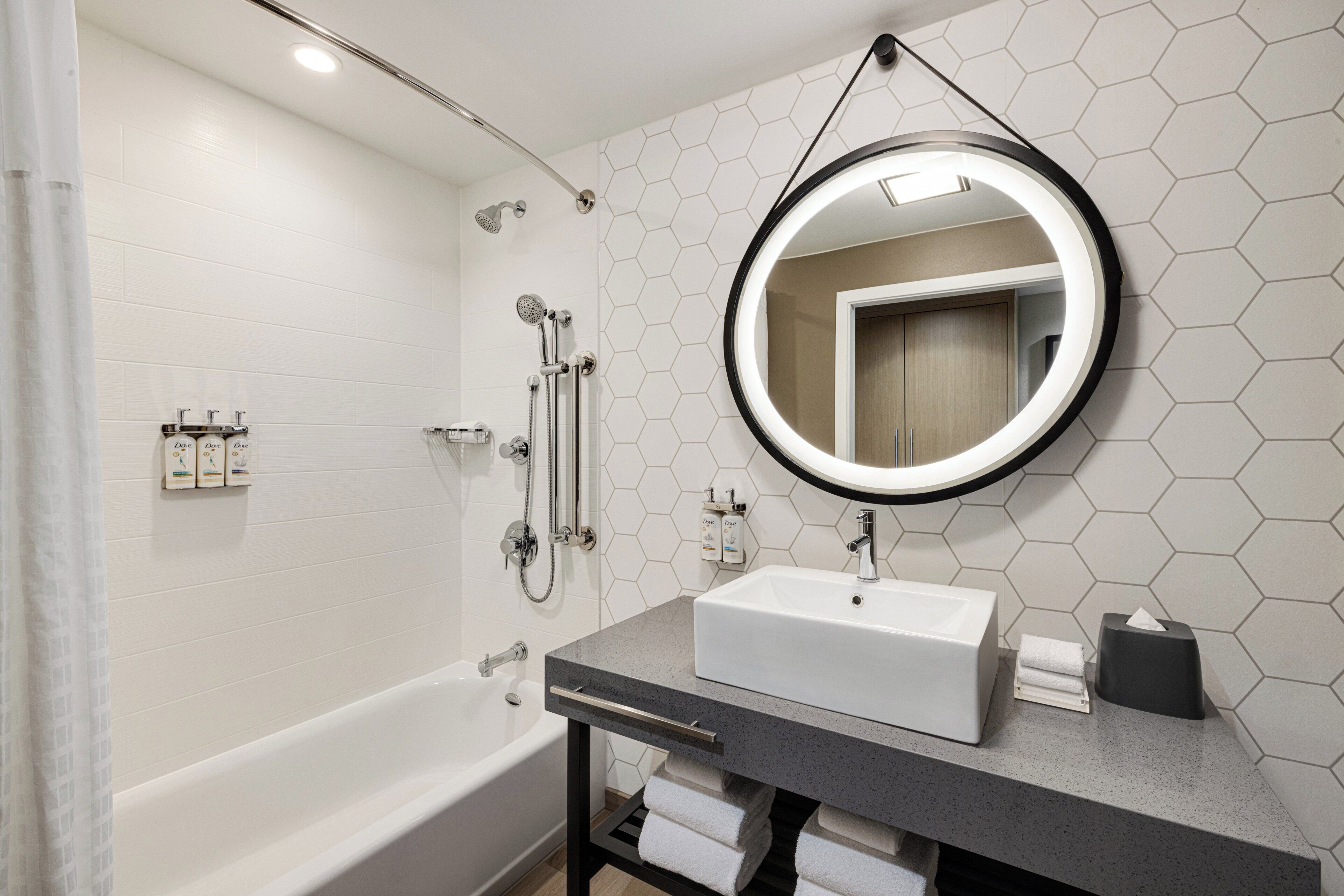 Un baño moderno con una bañera blanca, un espejo redondo y una encimera gris.