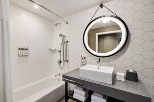 Un baño moderno con una bañera blanca, un espejo redondo y una encimera gris.