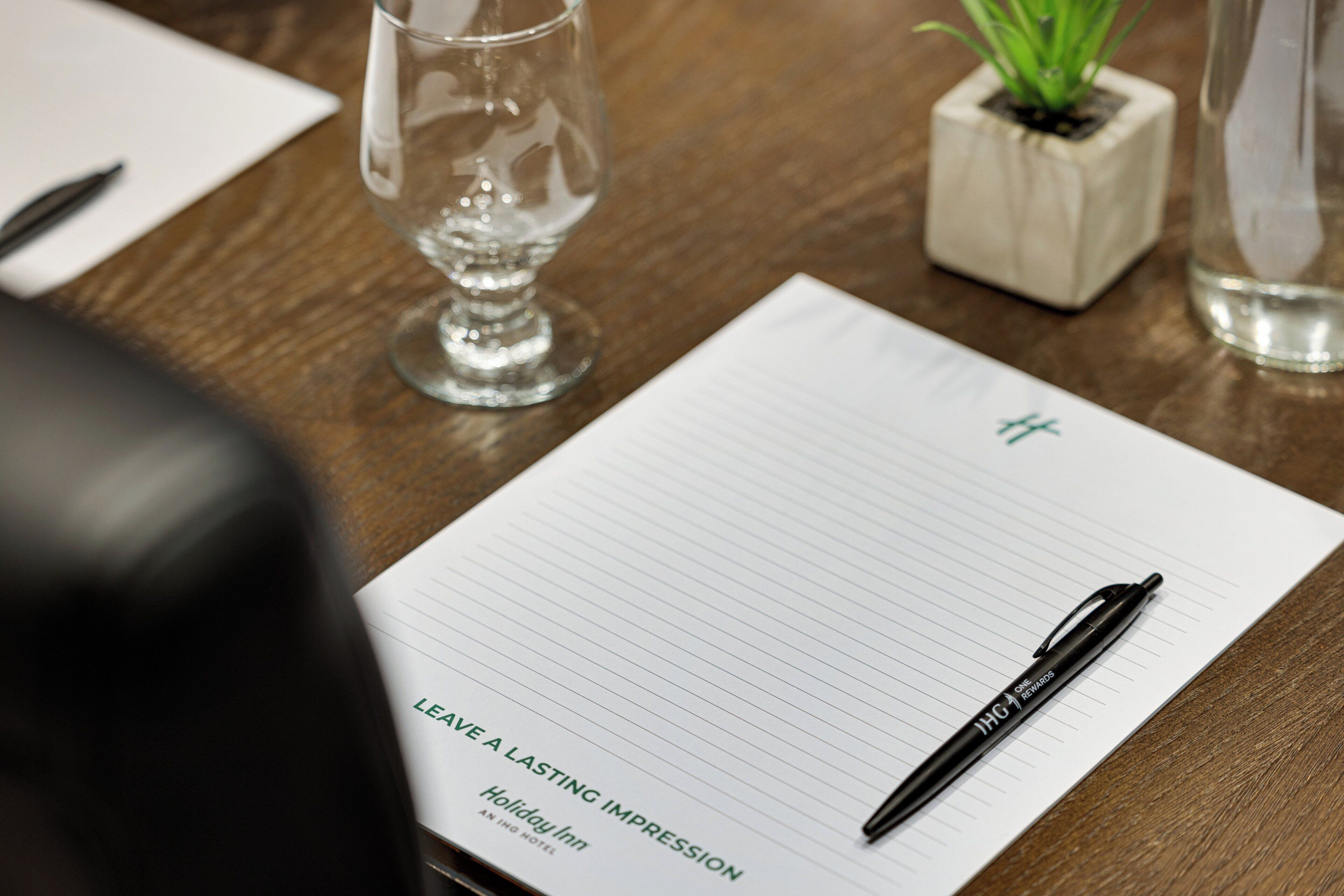A notebook with a pen on a wooden table, a glass of water, and a small plant in a cube planter.