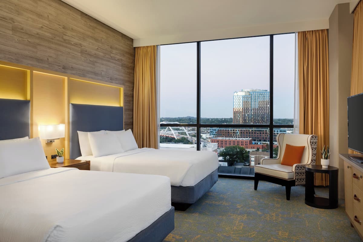 Habitación de hotel con dos camas, televisión, sillón y ventana grande con vista a la ciudad.