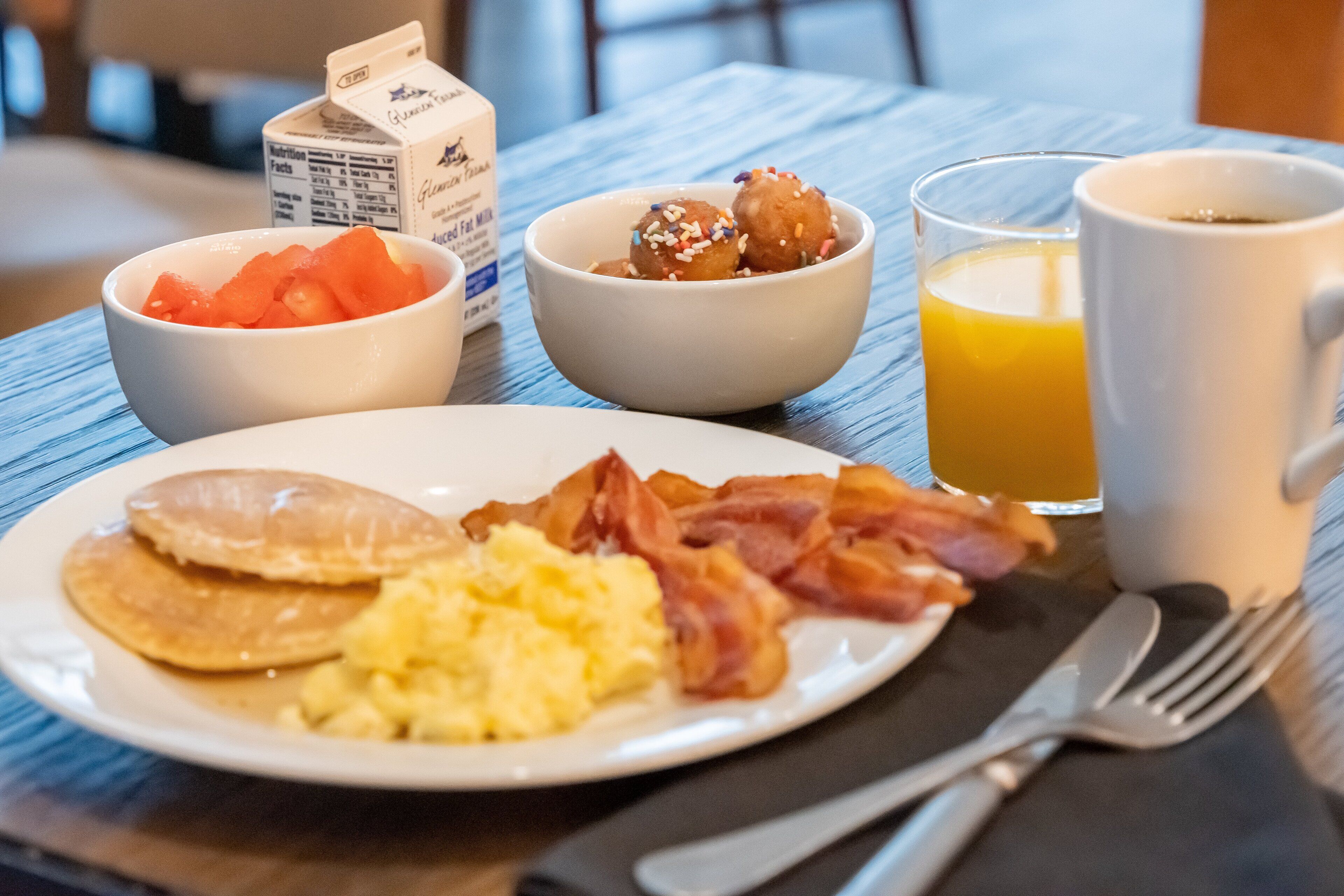 A breakfast plate with pancakes, bacon, scrambled eggs, and a bowl of fruit. There is a glass of orange juice and a white mug on the table.