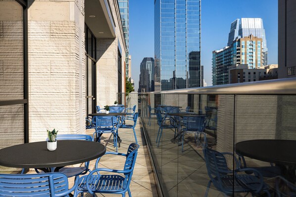 An outdoor terrace with blue metal chairs and tables, a potted plant, and a city skyline in the background.