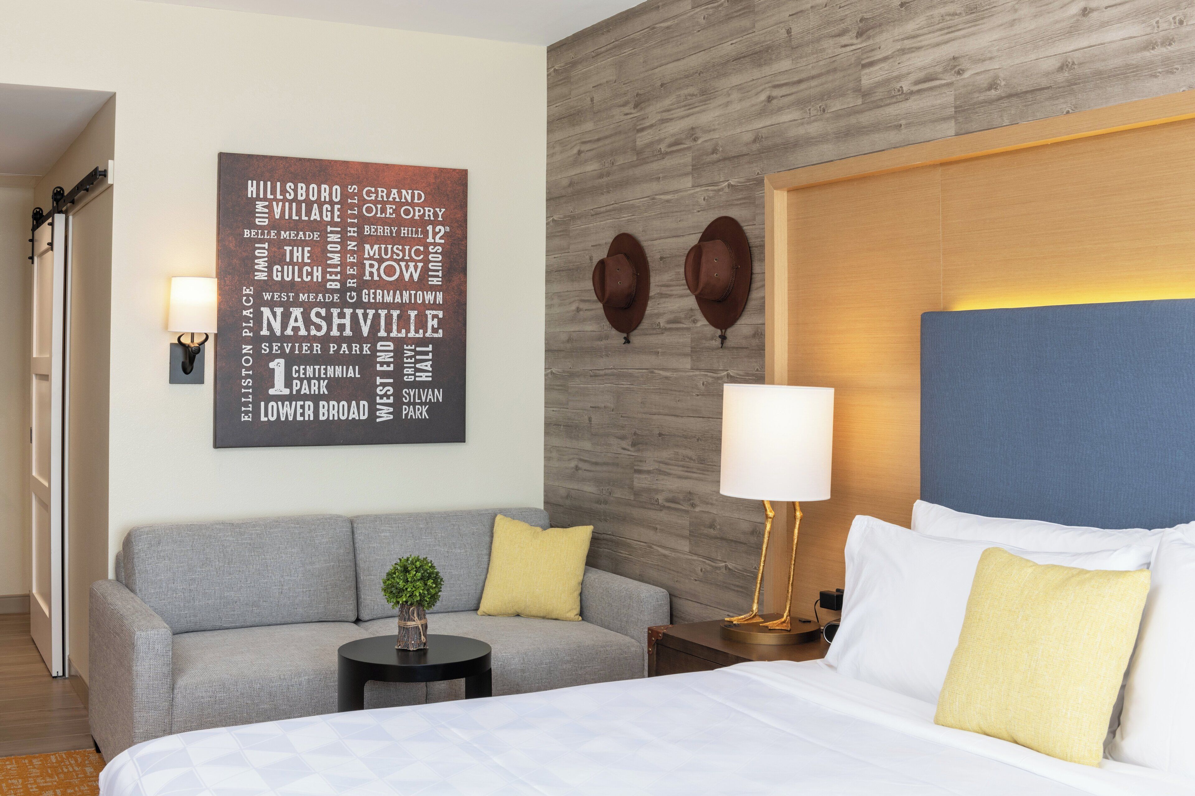 Ein Hotelzimmer mit einem Bett, einem grauen Sofa, einem kleinen Tisch mit einer Pflanze, einer Wand mit einer Wortwolke und zwei Hüten an der Wand.