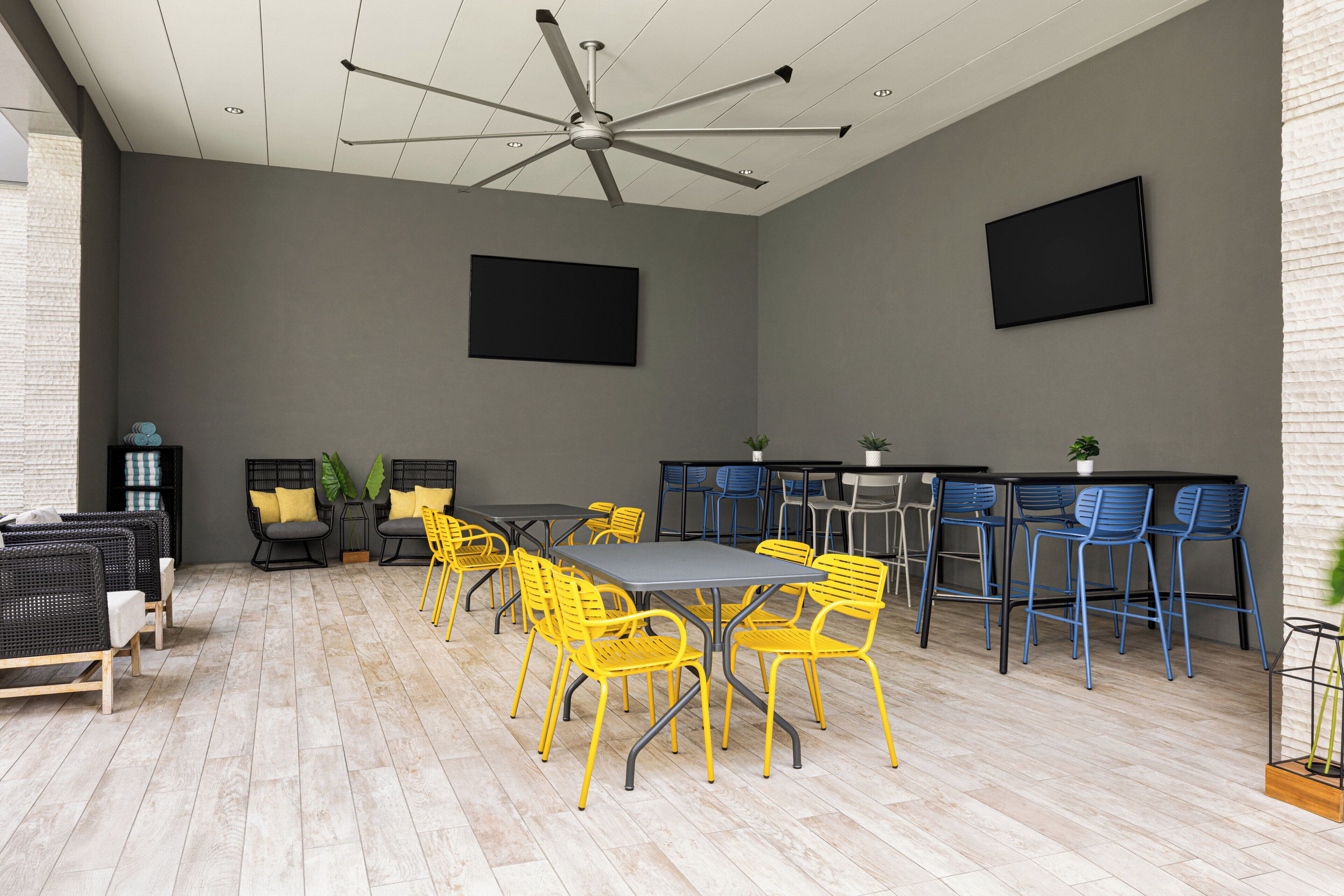 A modern meeting room with a central table, yellow and blue chairs, a ceiling fan, and two mounted screens.