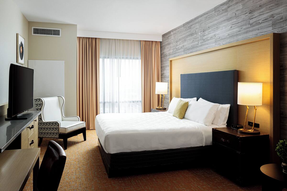Habitación de hotel con cama, escritorio con televisor, silla, mesita de noche con lámpara y ventana con cortinas.