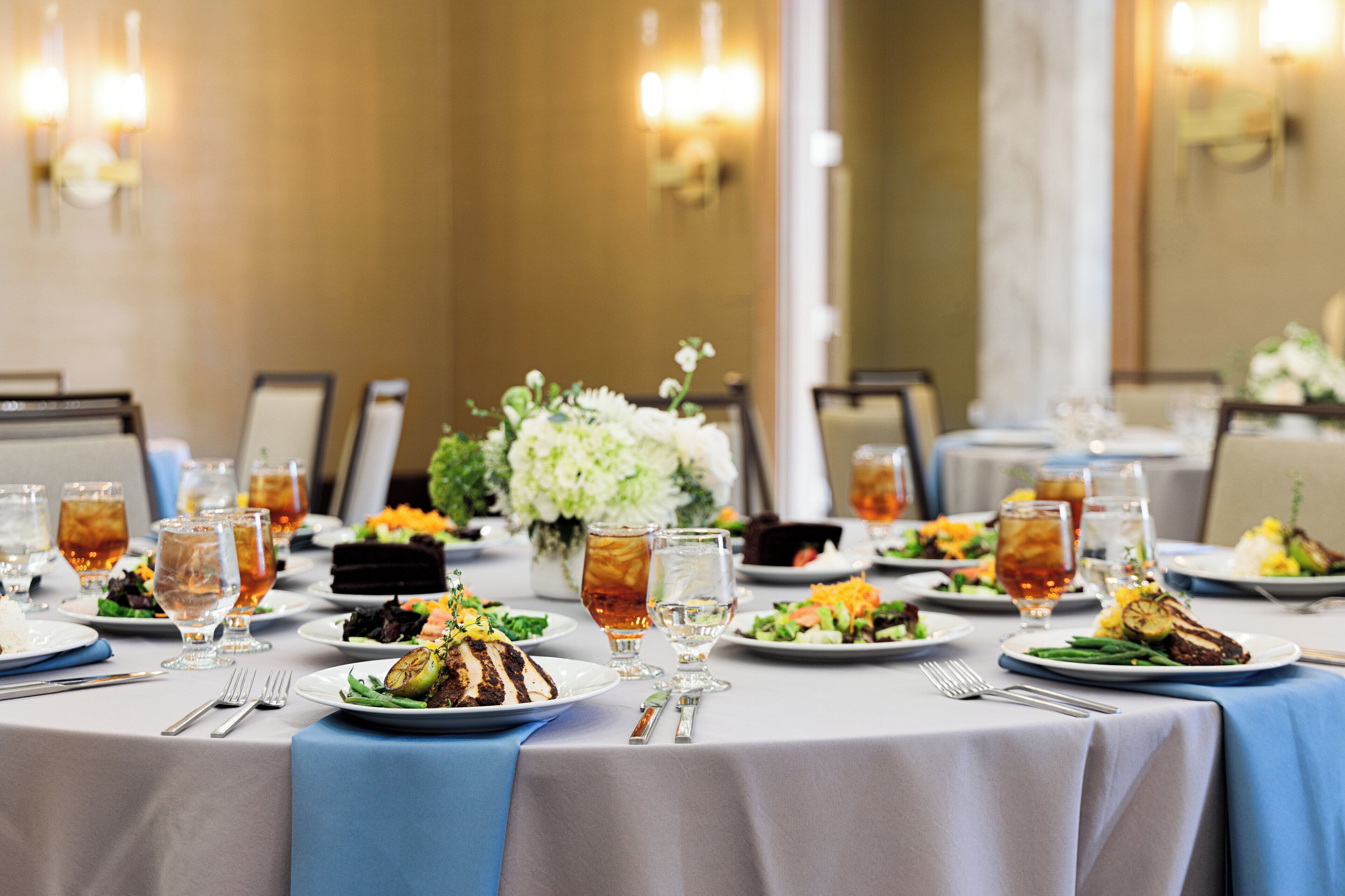 A dining table set with plates of food, glasses, and cutlery, with a blue table runner and floral centerpiece.