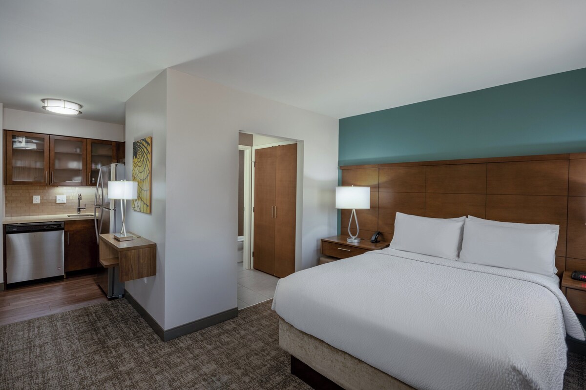 A hotel room with a bed, bedside table, lamp, and a view of the kitchen area.
