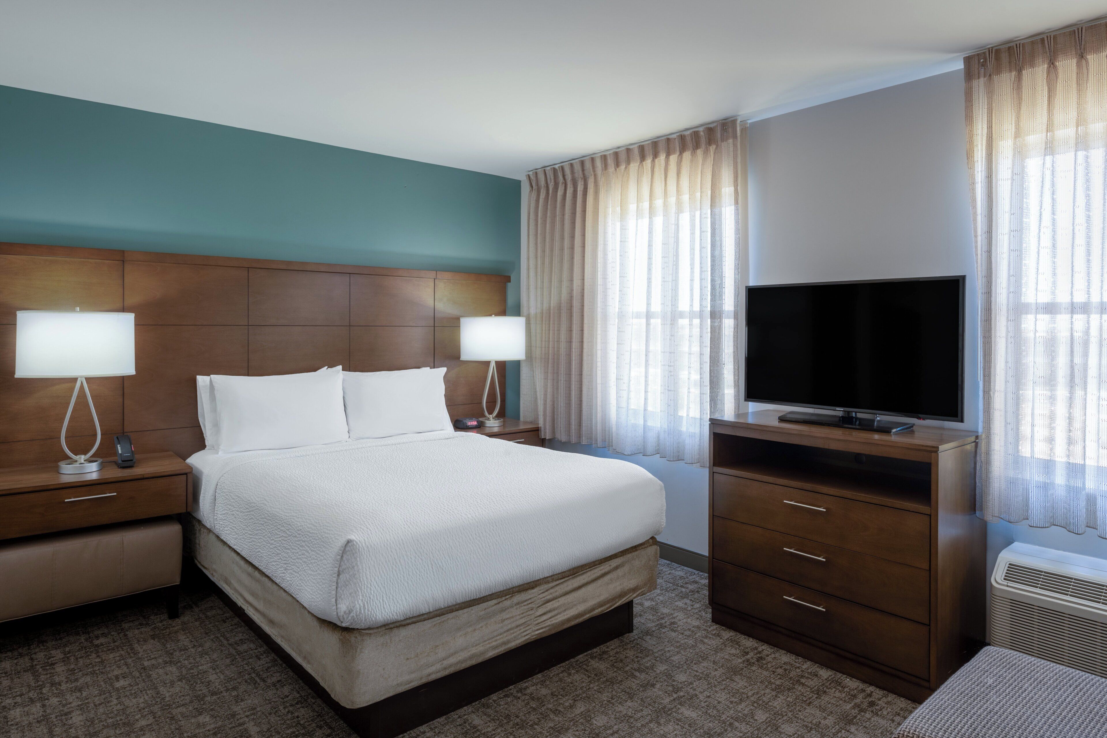 A hotel room with a large bed, two bedside tables with lamps, a TV on a stand, and a window with curtains.