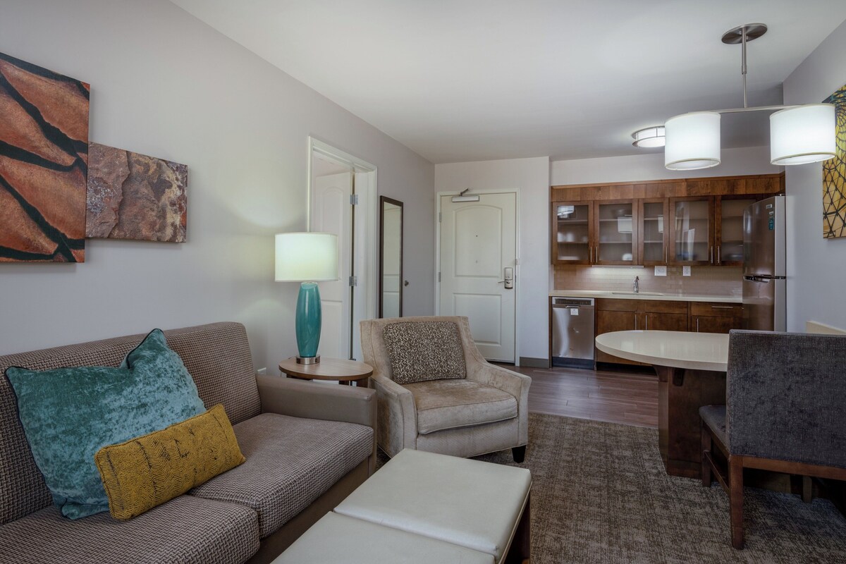 A living room with a sofa, armchair, and ottoman, a dining table with chairs, and a kitchen area with cabinets and a refrigerator.