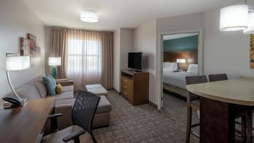 A hotel room with a sofa, a desk with a chair, a dining table with chairs, a TV, and a bed with a headboard.