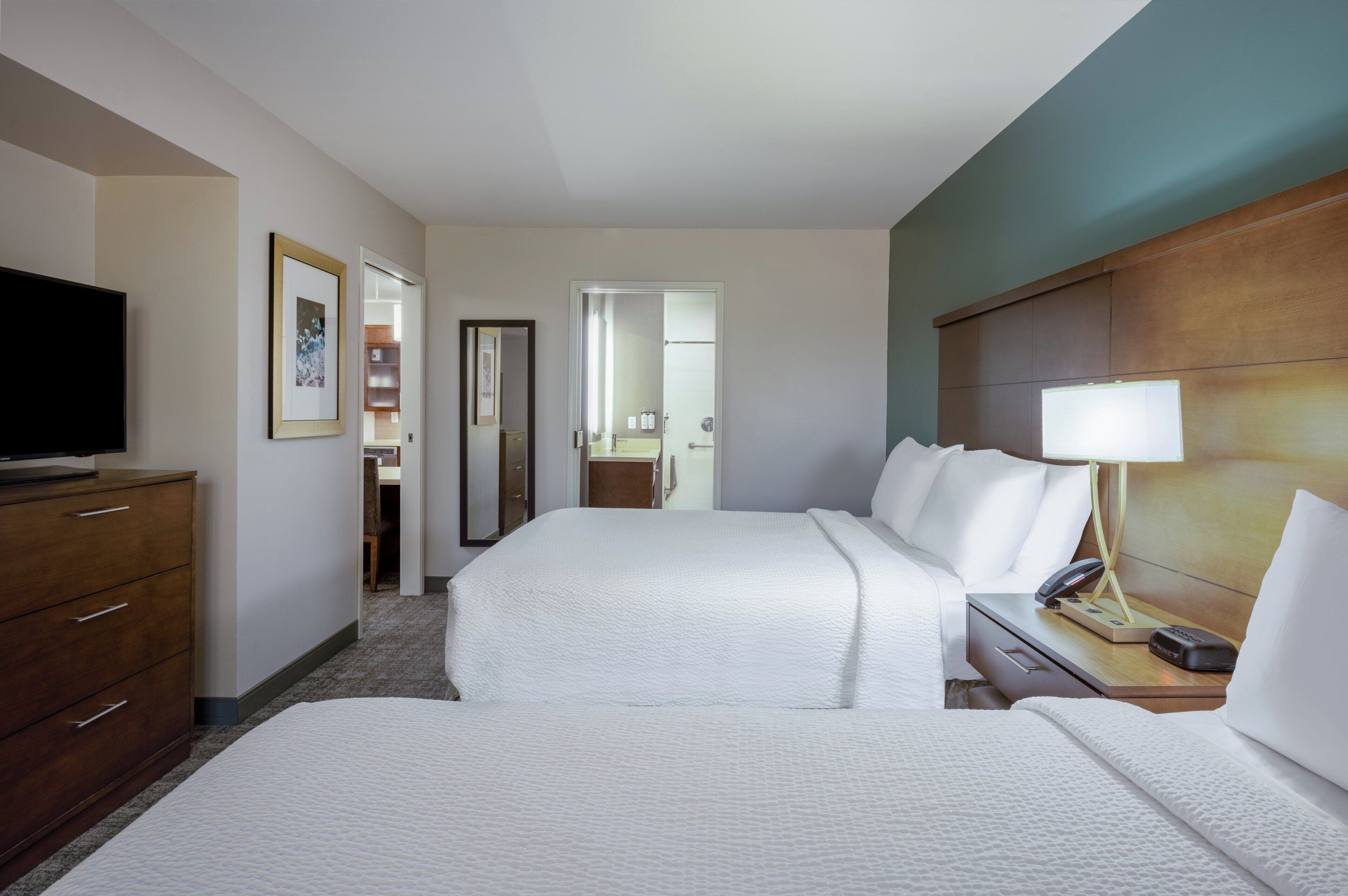 Una habitación de hotel con dos camas, televisión, mesita de noche con lámpara y cabecero de madera.