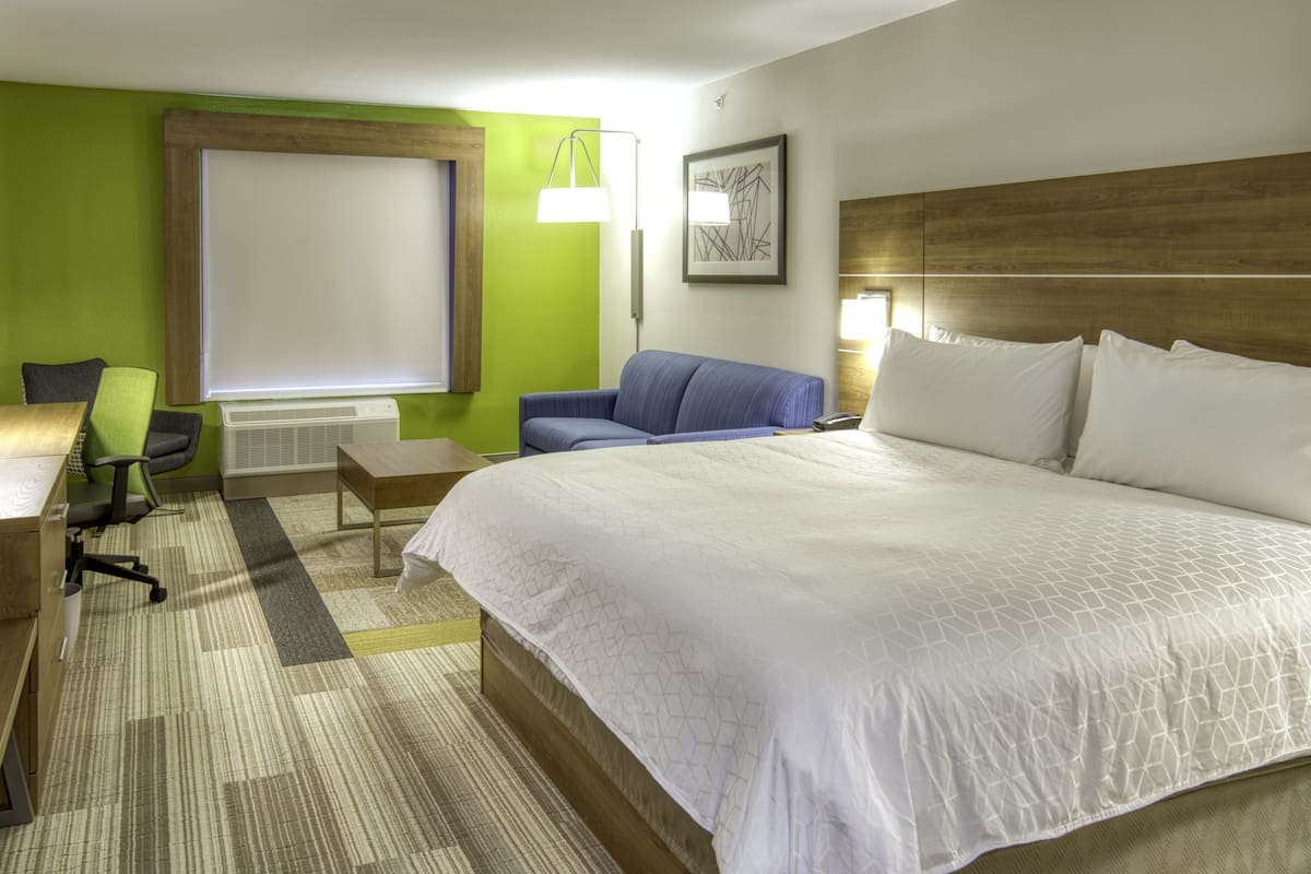 Een hotelkamer met een groot bed, een bureau met stoel, een staande lamp en een raam met jaloezieën.