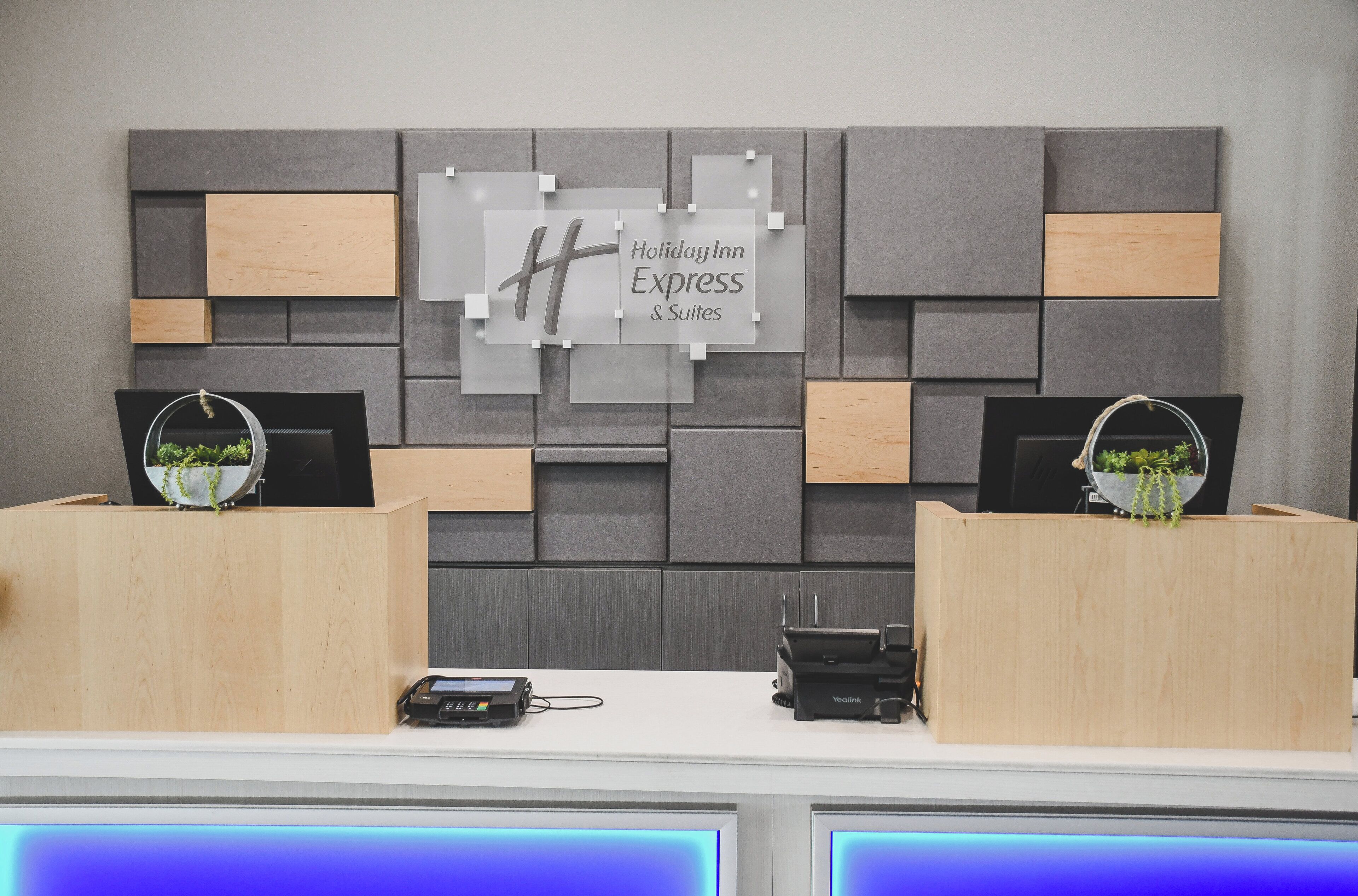 A reception desk with a modern design, featuring a grey and wooden panel wall, a sign for Holiday Inn Express & Suites, and two potted plants.