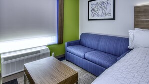 A hotel room with a large window, a blue sofa, a wooden table, and a bed with white bedding.