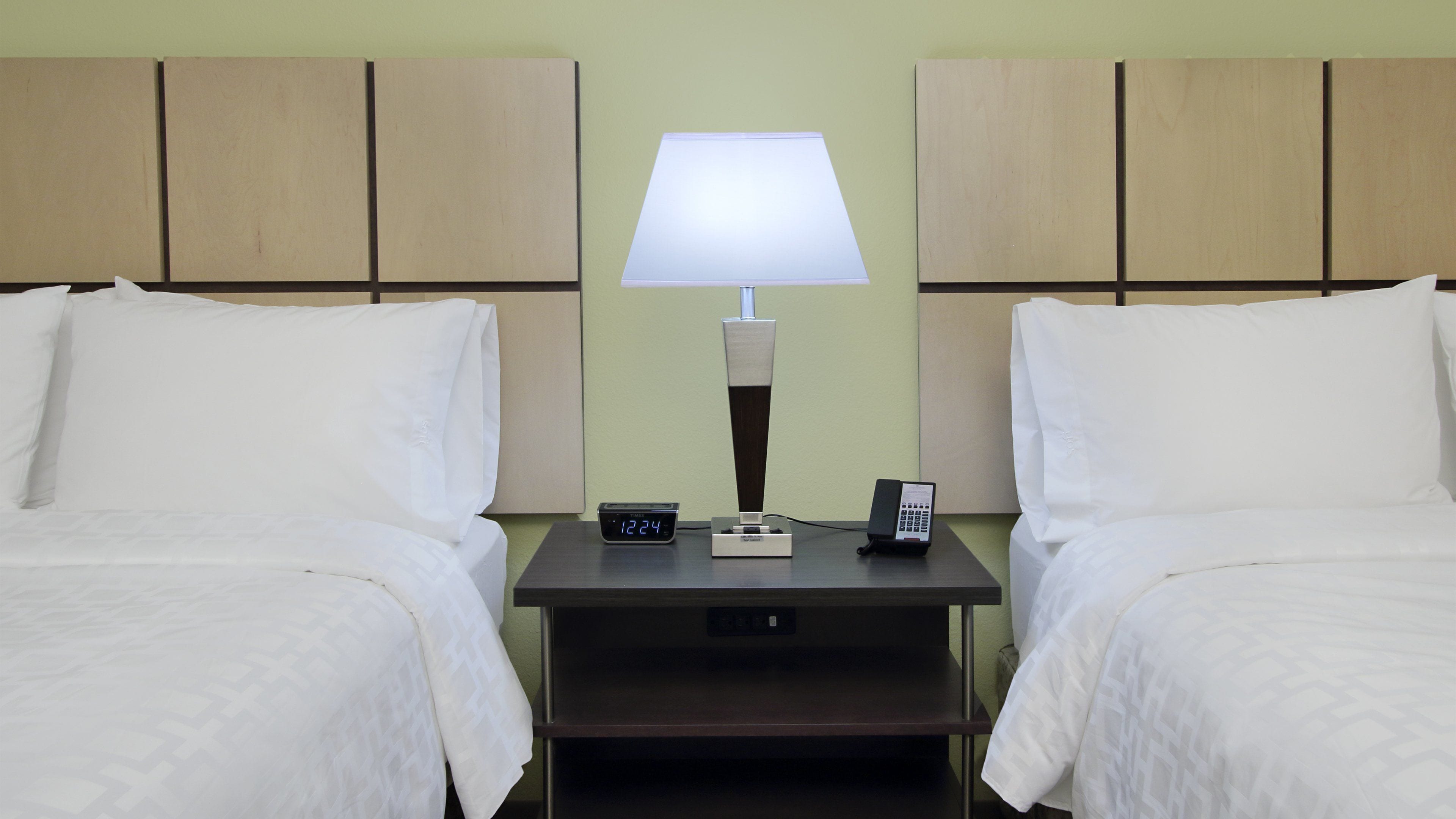 Habitación de hotel con dos camas, una mesita de noche con lámpara, un reloj digital y un teléfono.