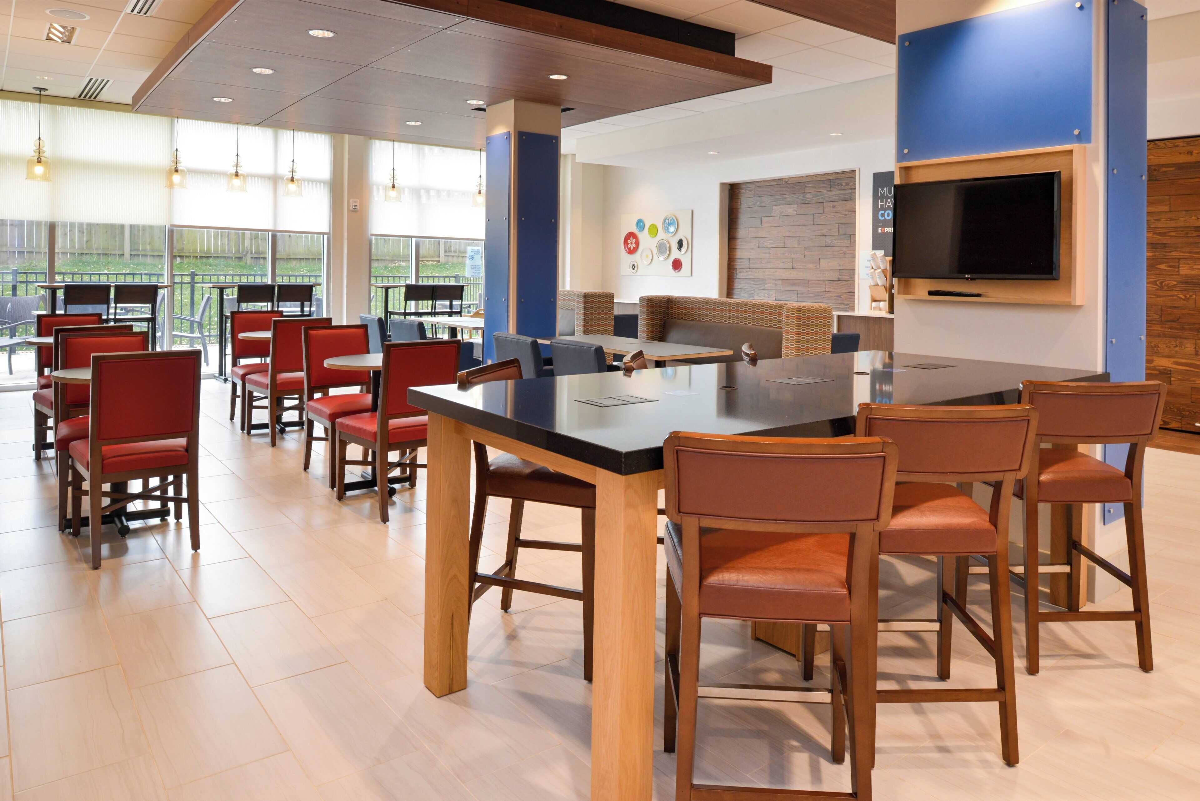 A modern dining area with a long table and multiple chairs, a flat-screen TV mounted on the wall, and a view of the outdoors through large windows.