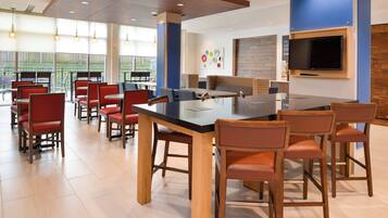 A modern dining area with a long table and multiple chairs, a flat-screen TV mounted on the wall, and a view of the outdoors through large windows.