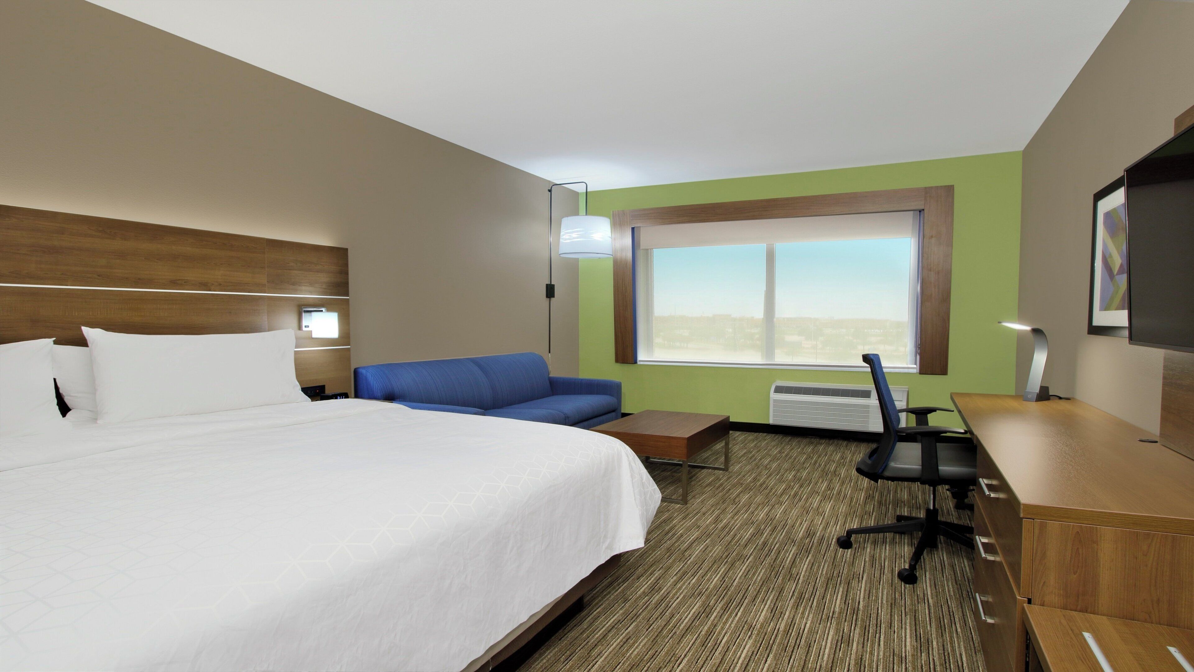 Hotelkamer met een bed, een blauwe bank, een bureau met stoel, een raam en een airconditioning.