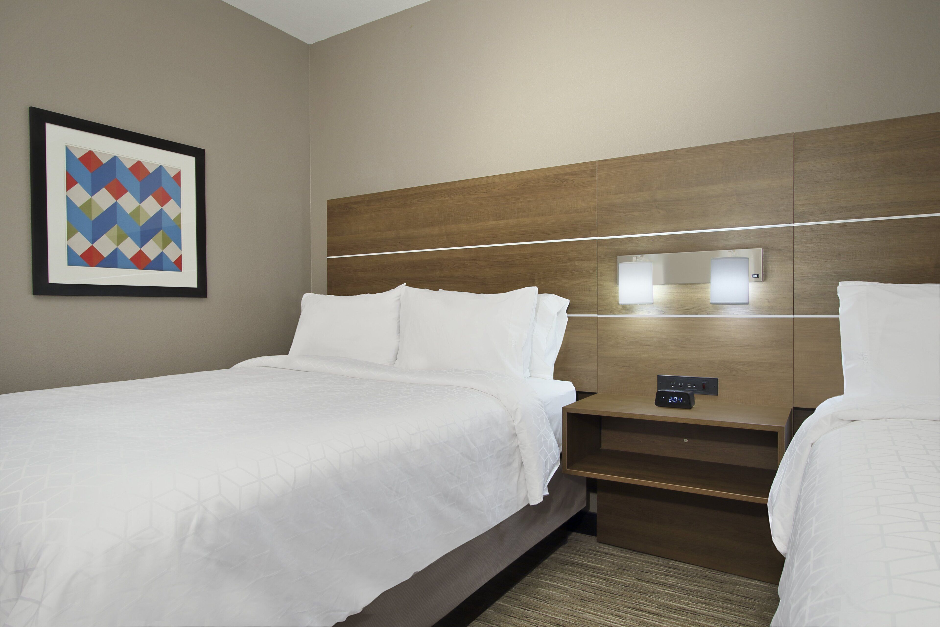 A hotel room with two beds, a wooden headboard, a nightstand with a clock, and a framed geometric art piece on the wall.