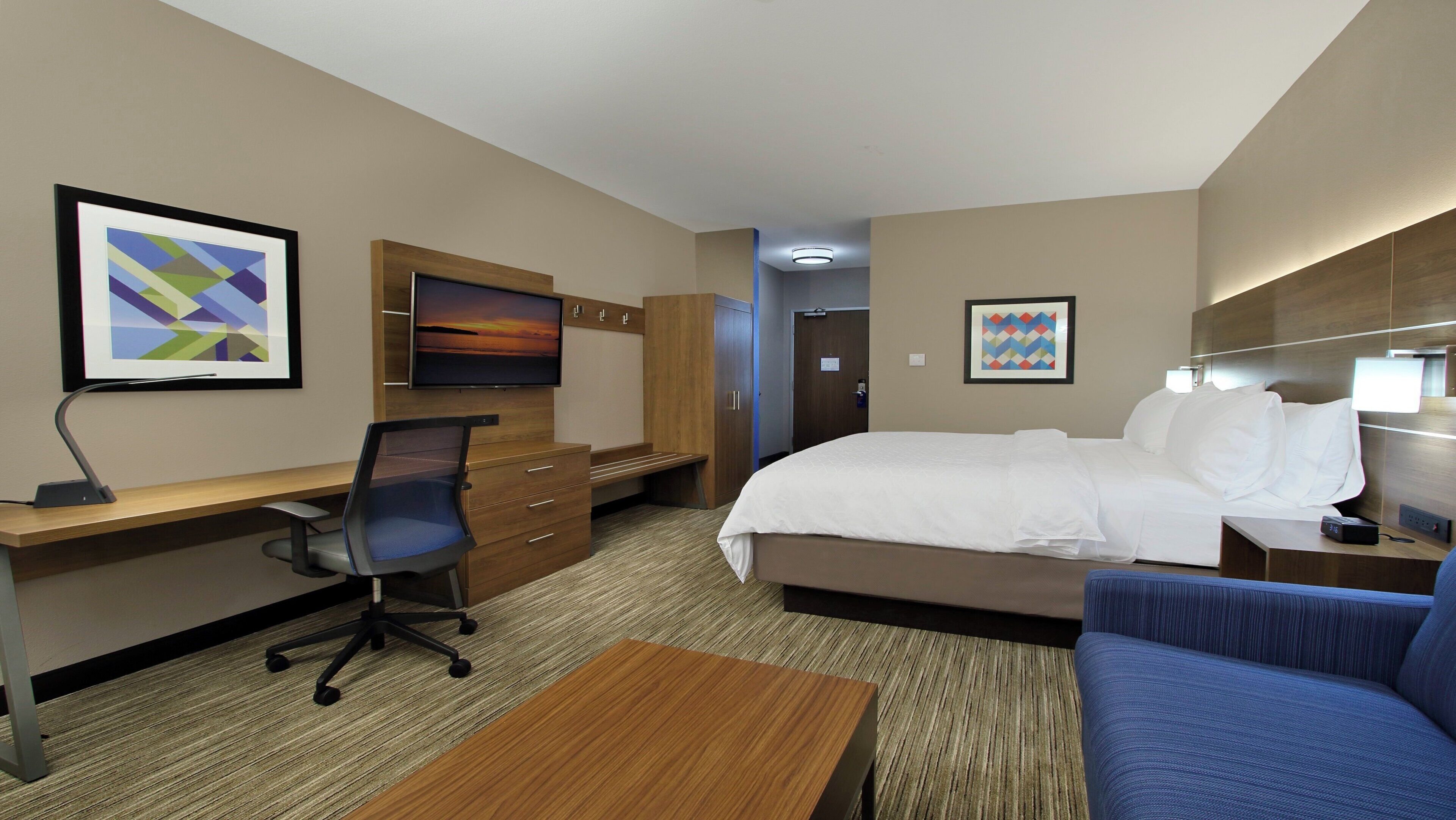A hotel room with a bed, a desk with a computer, a chair, a TV, and artwork on the walls.