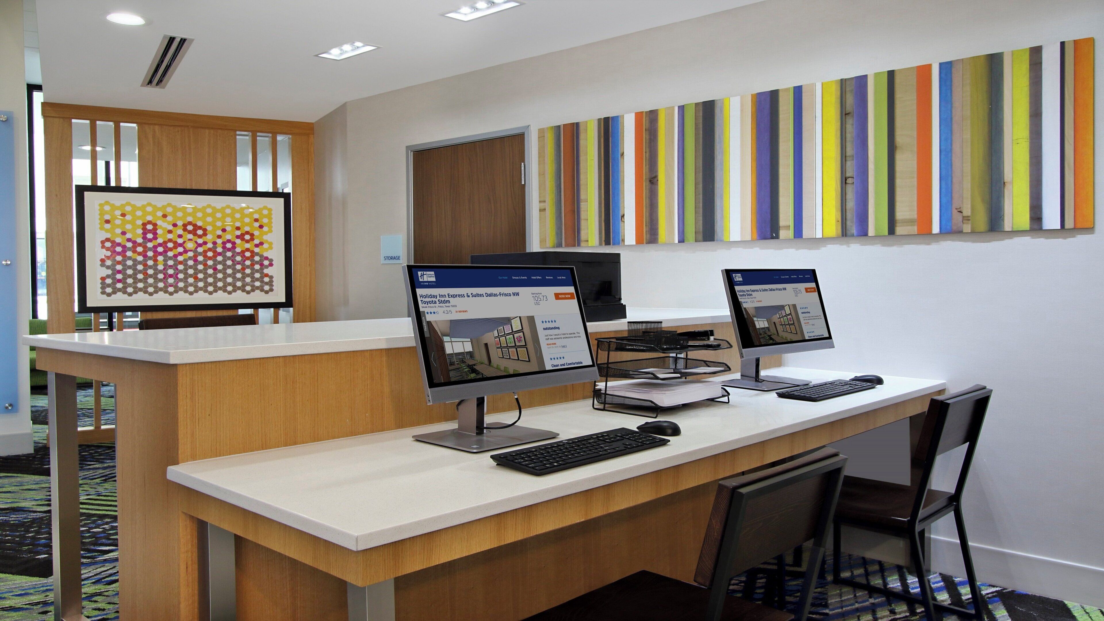 A reception desk with two monitors, a keyboard, and a chair. There is a colorful artwork on the wall and a storage area with a sign.