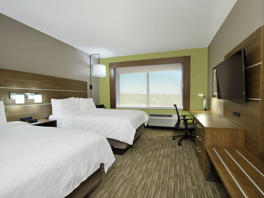 A hotel room with two beds, a desk, a chair, a flat-screen TV, and a large window offering a city view.