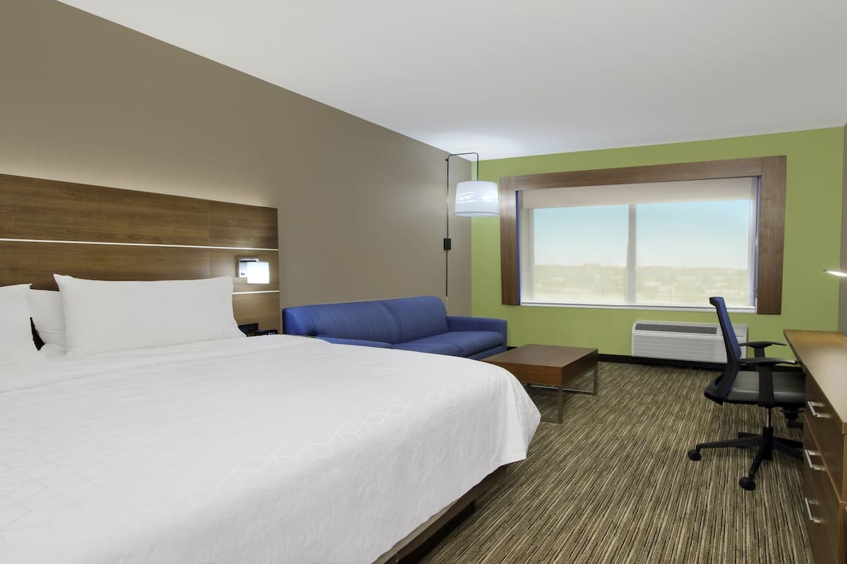 A hotel room with a large bed, a blue sofa, a desk with a chair, a window with a view, and a mounted TV.