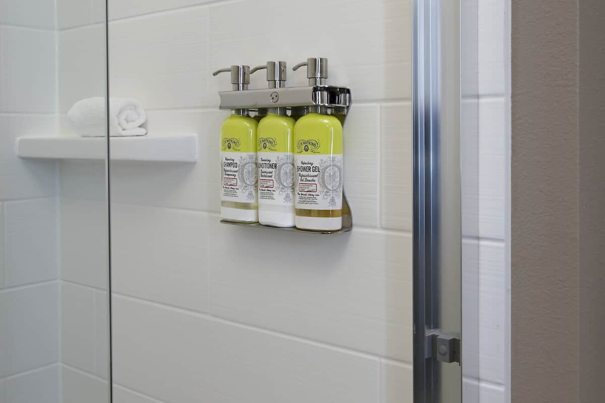 A bathroom with a white tiled wall, a glass shower door, and a shelf holding three bottles of shampoo and conditioner.
