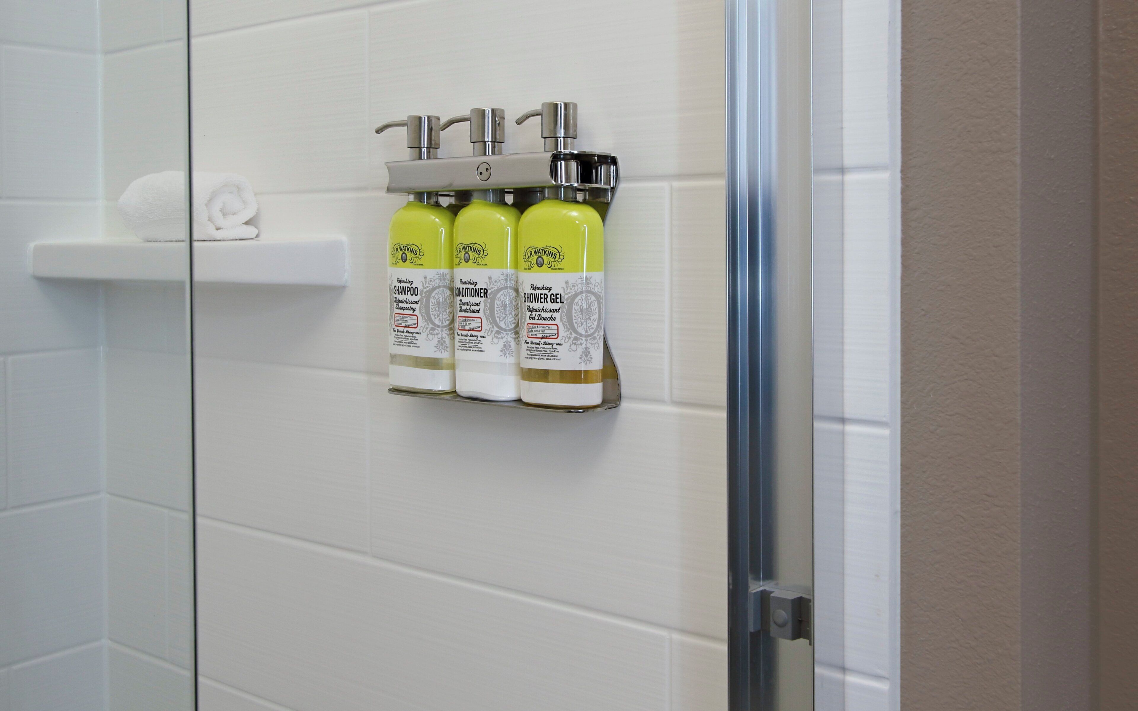 Et badeværelse med hvide flisemursten, en brusegøgl af glas og en hylde med tre shampoo- og conditionerflasker.