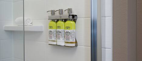 A bathroom with white tiled walls, a glass shower door, and a shelf holding three bottles of shampoo and conditioner.