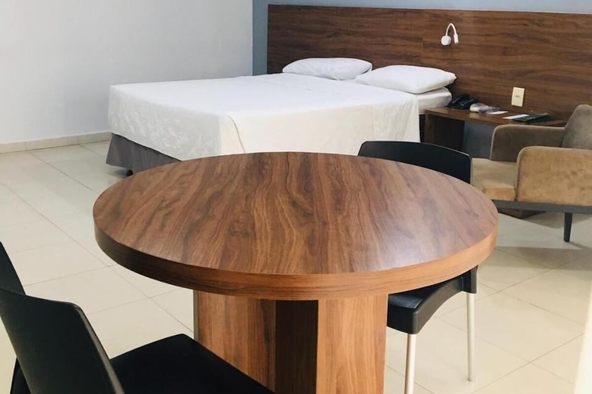 A hotel room with a round wooden table, two black chairs, a bed with white bedding, a nightstand with a lamp, and a headboard.