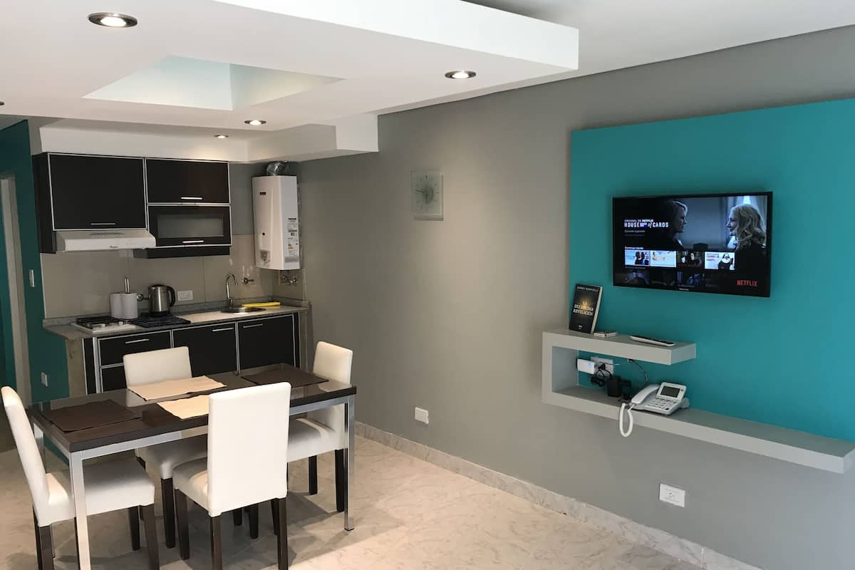 A modern kitchen with dark cabinets, a microwave, and a sink. There is a dining area with a table and chairs. A TV mounted on the wall displays a show.