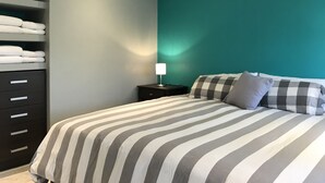 Cama bem arrumada com roupa de cama listrada, mesa de cabeceira com abajur e um armário com toalhas dobradas.