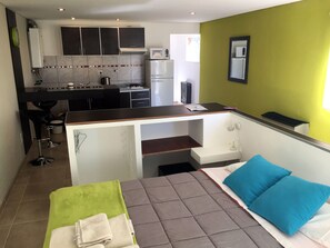 Une chambre d’hôtel compacte comprenant un lit, une serviette verte, un oreiller bleu, une kitchenette et un petit bureau.