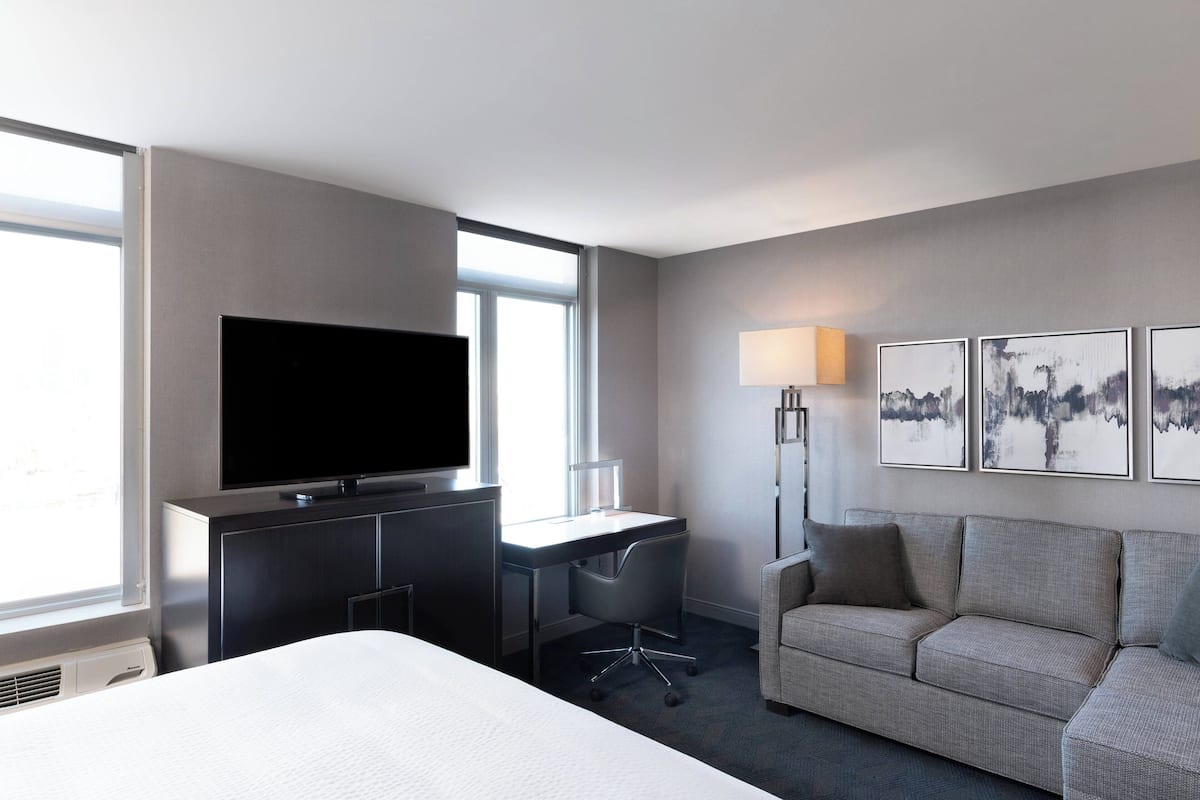 A hotel room with a bed, a desk with a chair, a television, a sofa, and artwork on the wall.