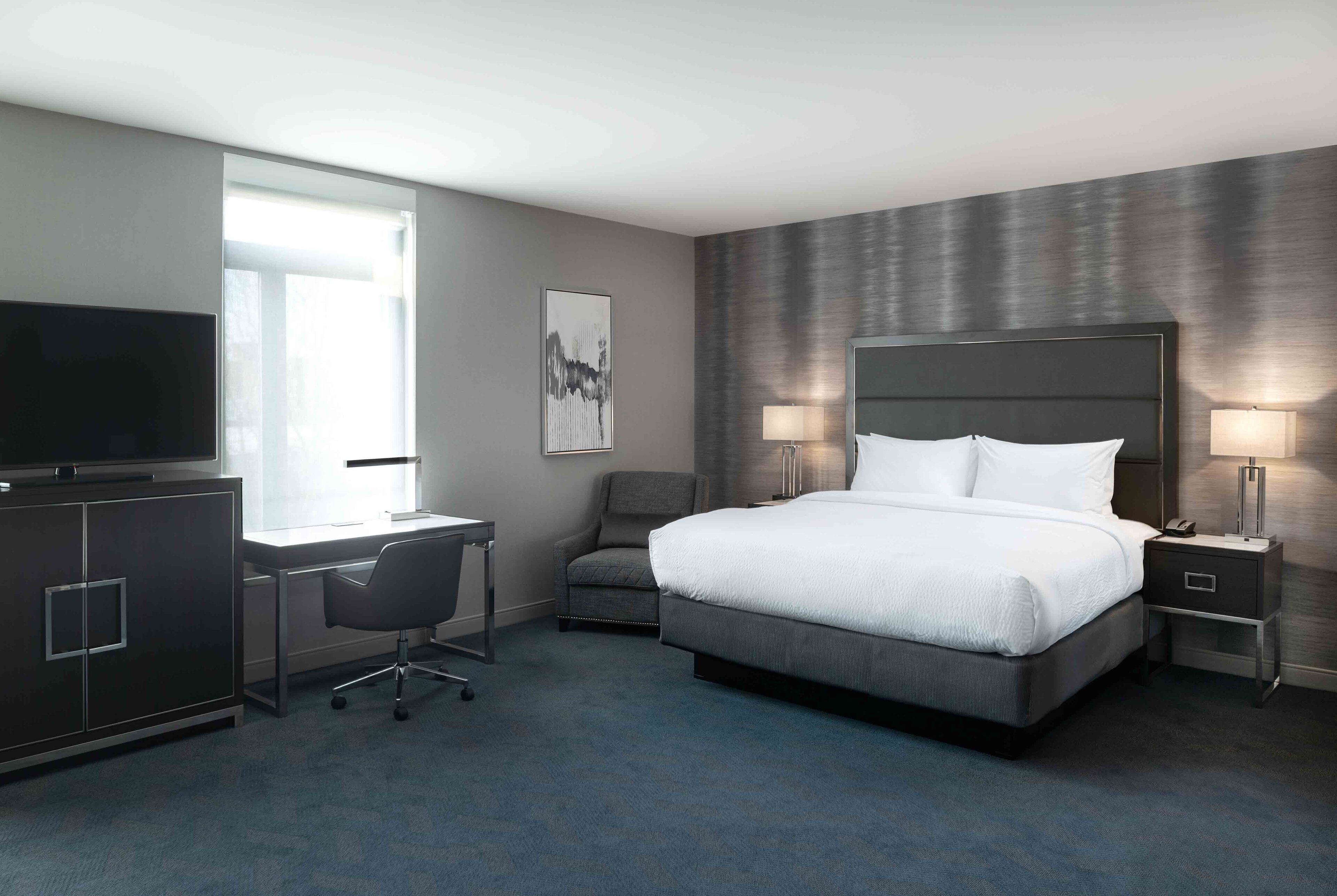 A hotel room with a large bed, bedside tables with lamps, a desk with a chair, a television, and a window.