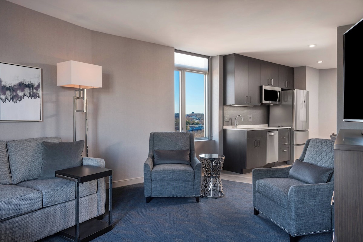 A modern hotel room with a grey sofa, two armchairs, a small table, a floor lamp, and a view of the city through a large window.