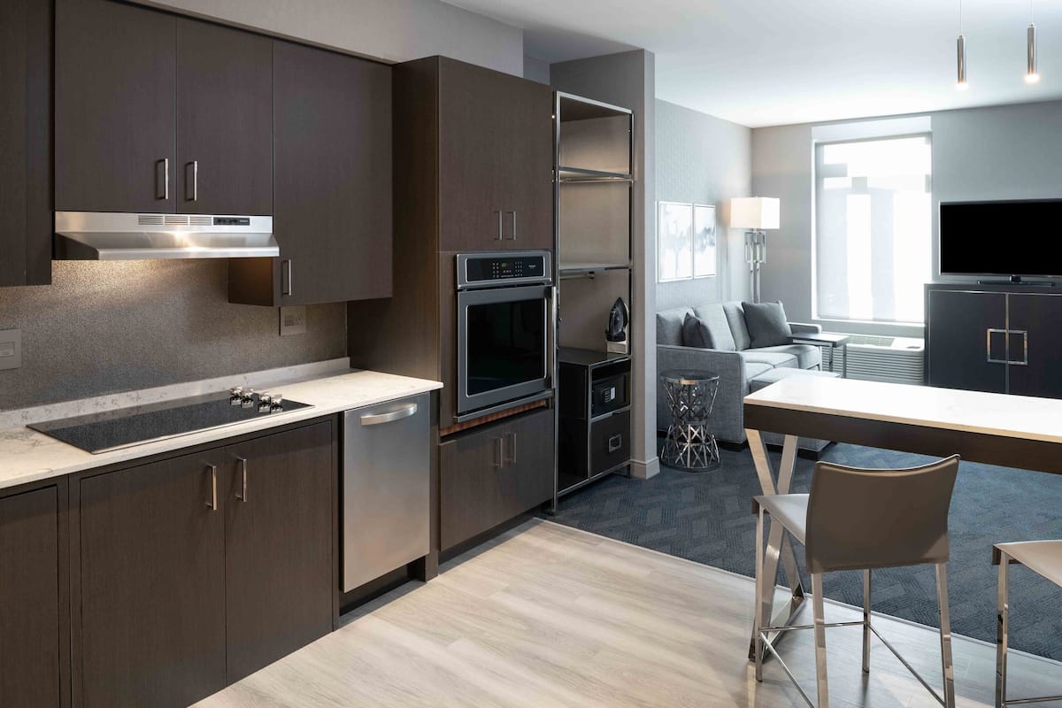 A modern kitchen with dark cabinetry, a built-in oven, and a microwave. There is a dining area with a table and chairs, and a TV mounted on the wall.