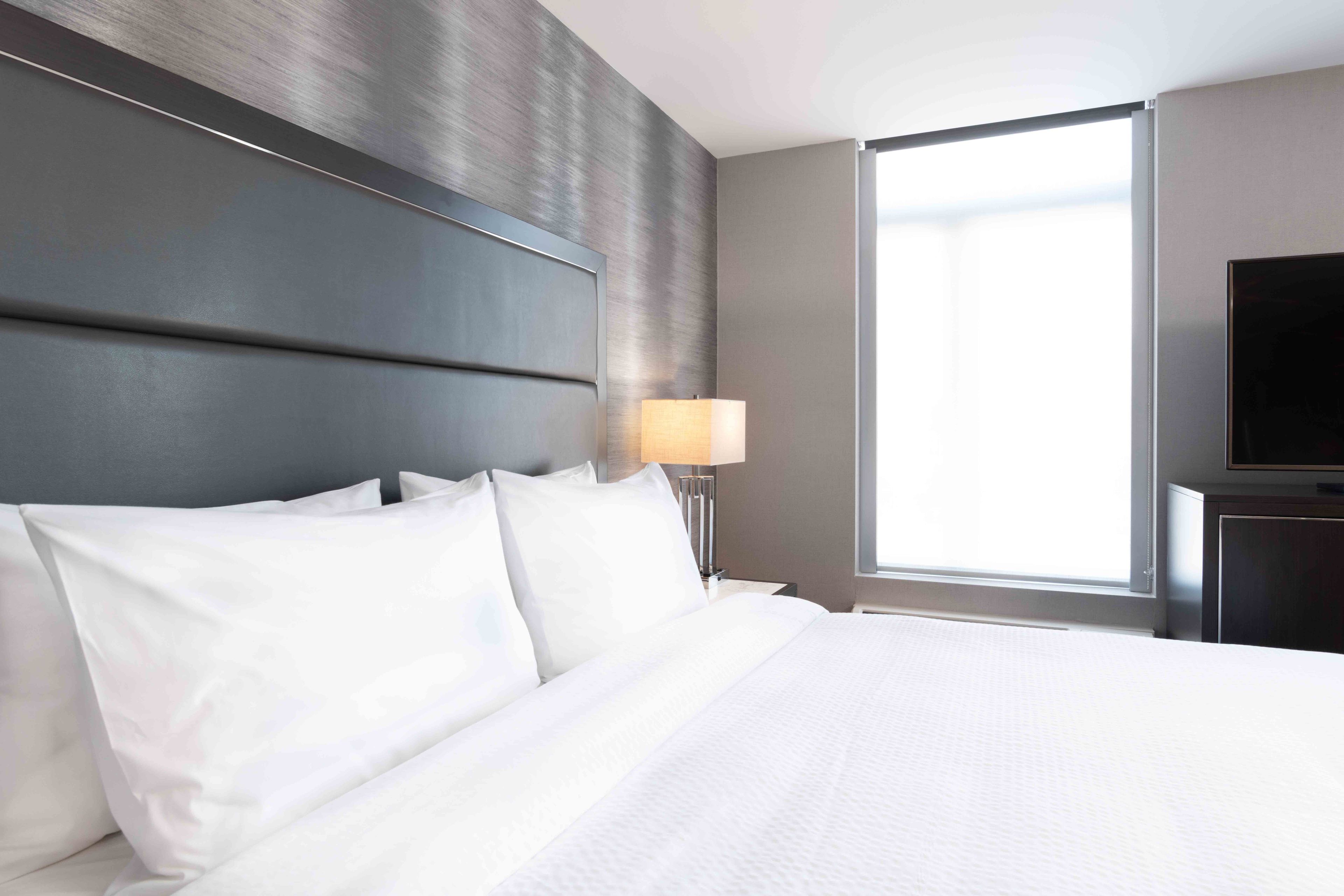 Quarto de hotel com uma cama grande, televisão de tela plana, janela com cortinas e abajur em uma mesa de cabeceira.