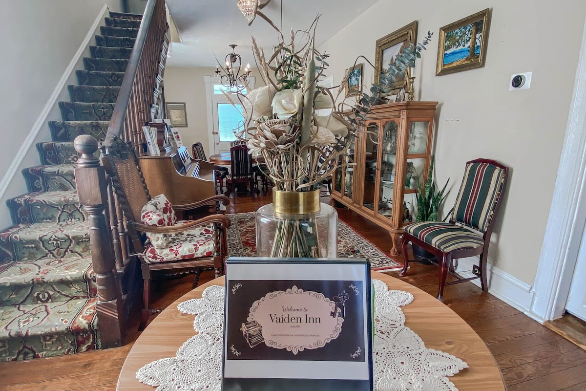 A reception area with a wooden staircase, a chandelier, a floral arrangement, and a welcome sign for the Vaiden Inn.