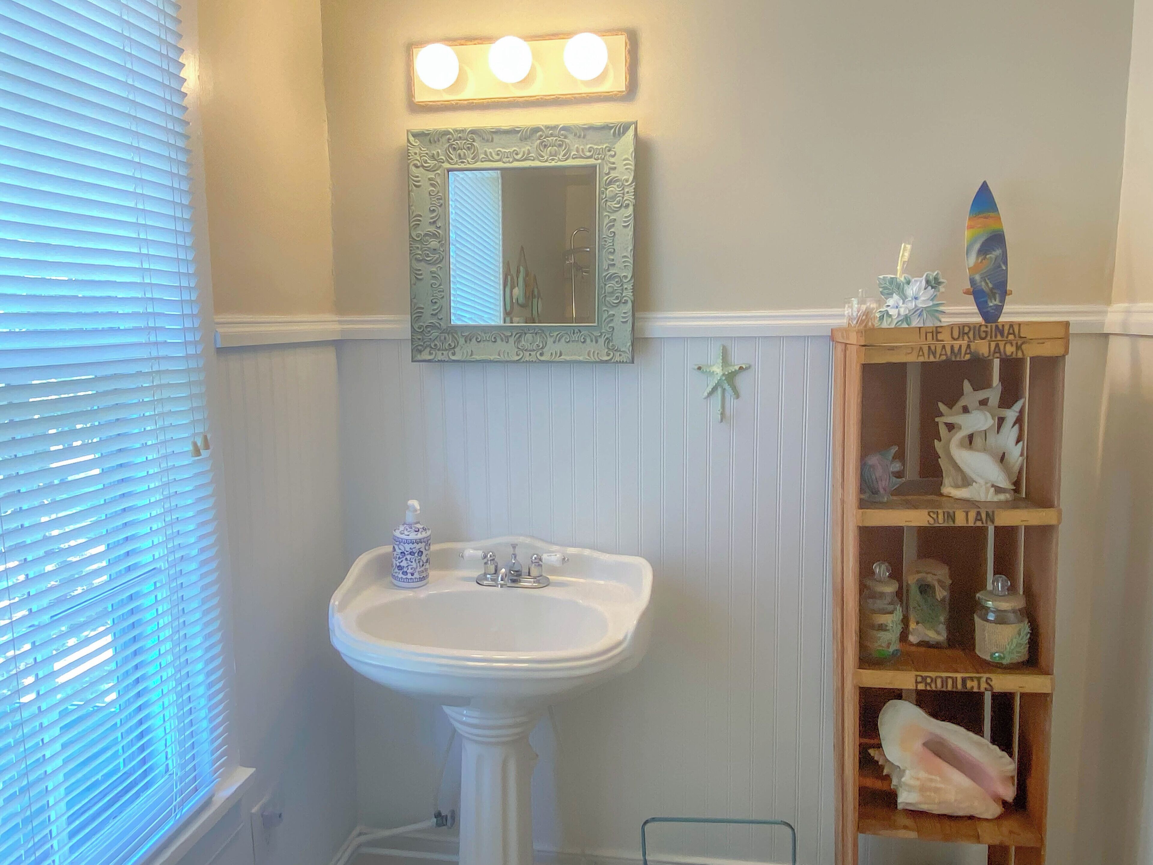 Une salle de bain avec un lavabo sur colonne, un miroir de coiffeuse et une étagère ornée d’objets décoratifs.