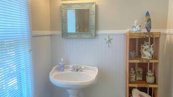 Une salle de bain avec un lavabo sur colonne, un miroir de coiffeuse et une étagère ornée d’objets décoratifs.