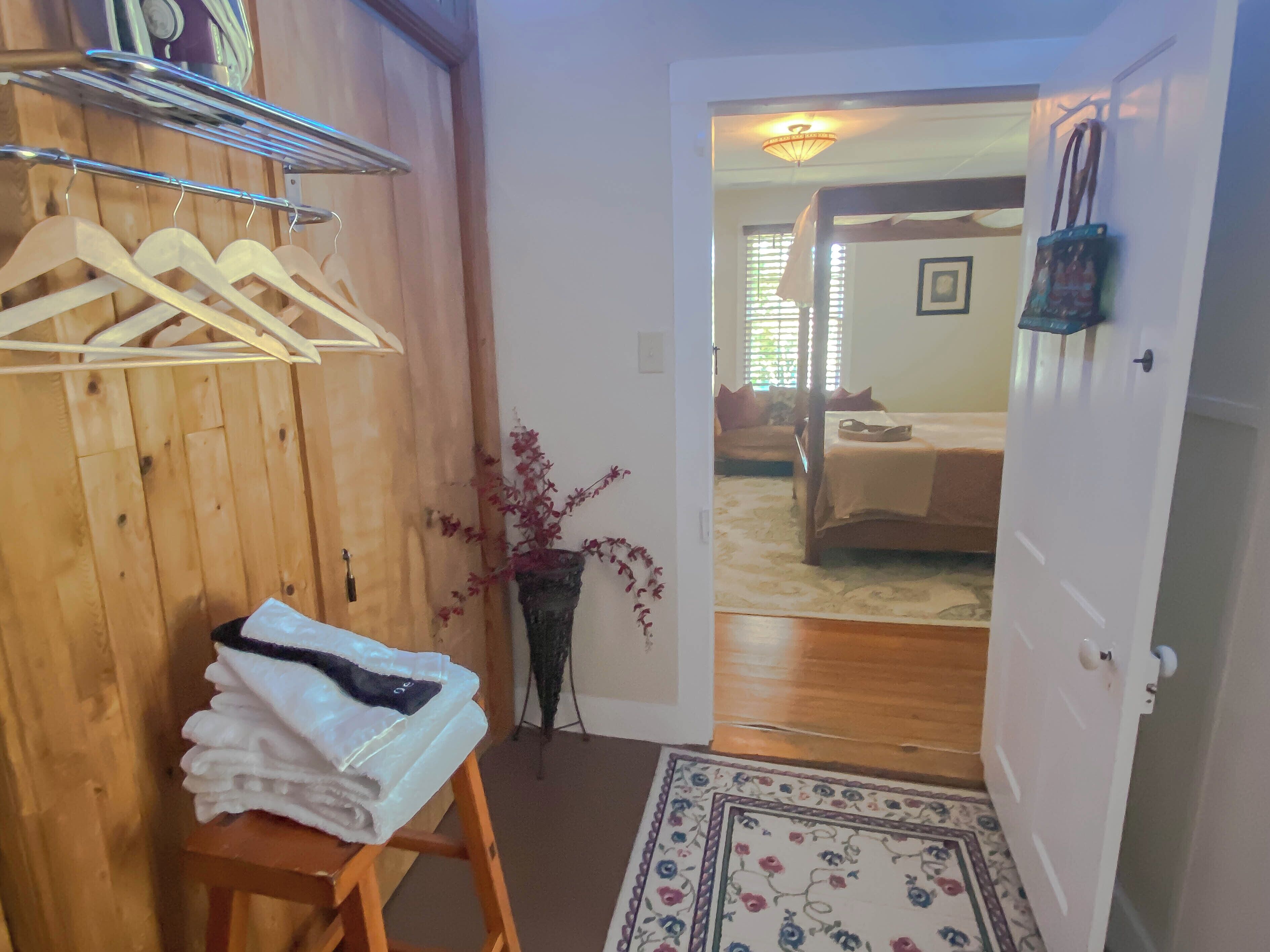 A room with wooden walls, a bed with a headboard, a small table with folded towels, and a door leading to another room.