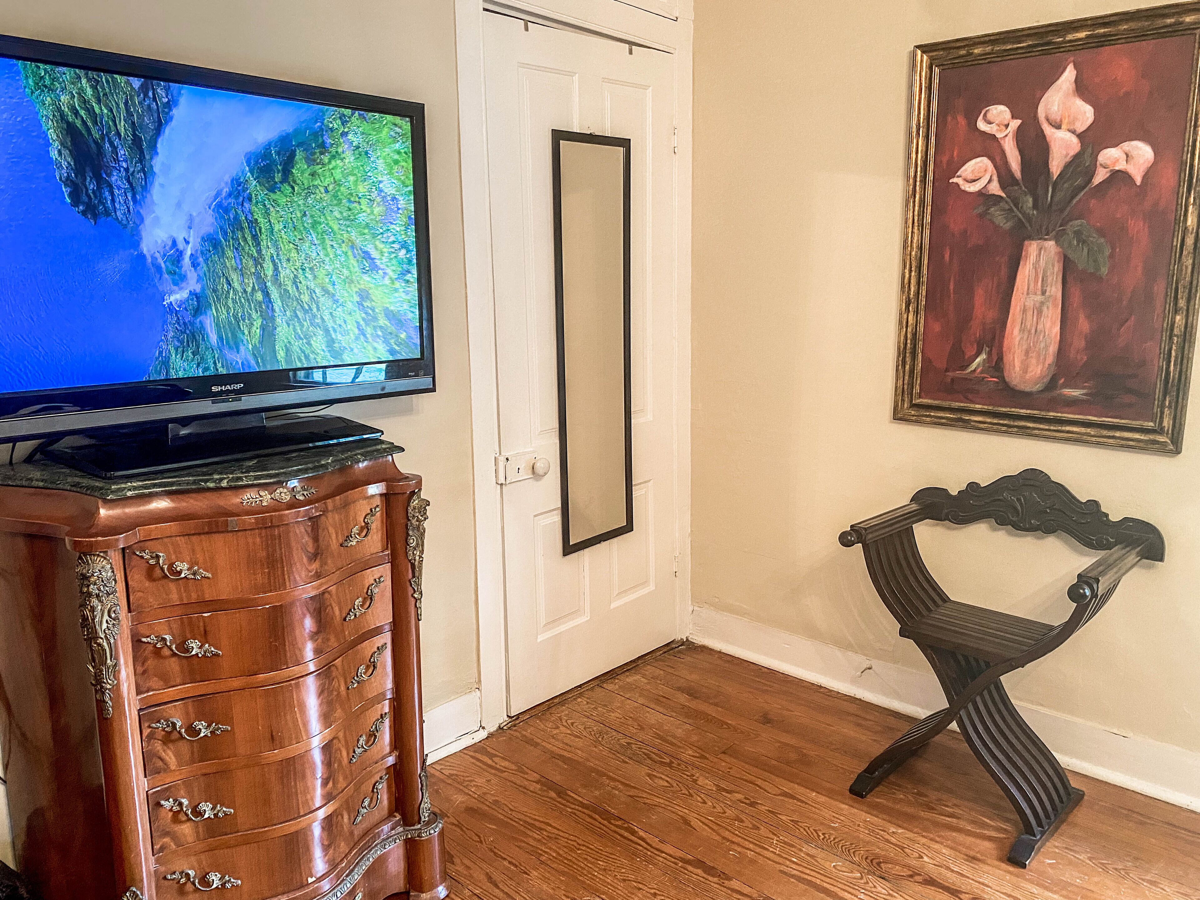 Ein Zimmer mit einem hölzernen Schminktisch, einem Fernseher darauf, einer Tür und einem gerahmten Gemälde an der Wand.