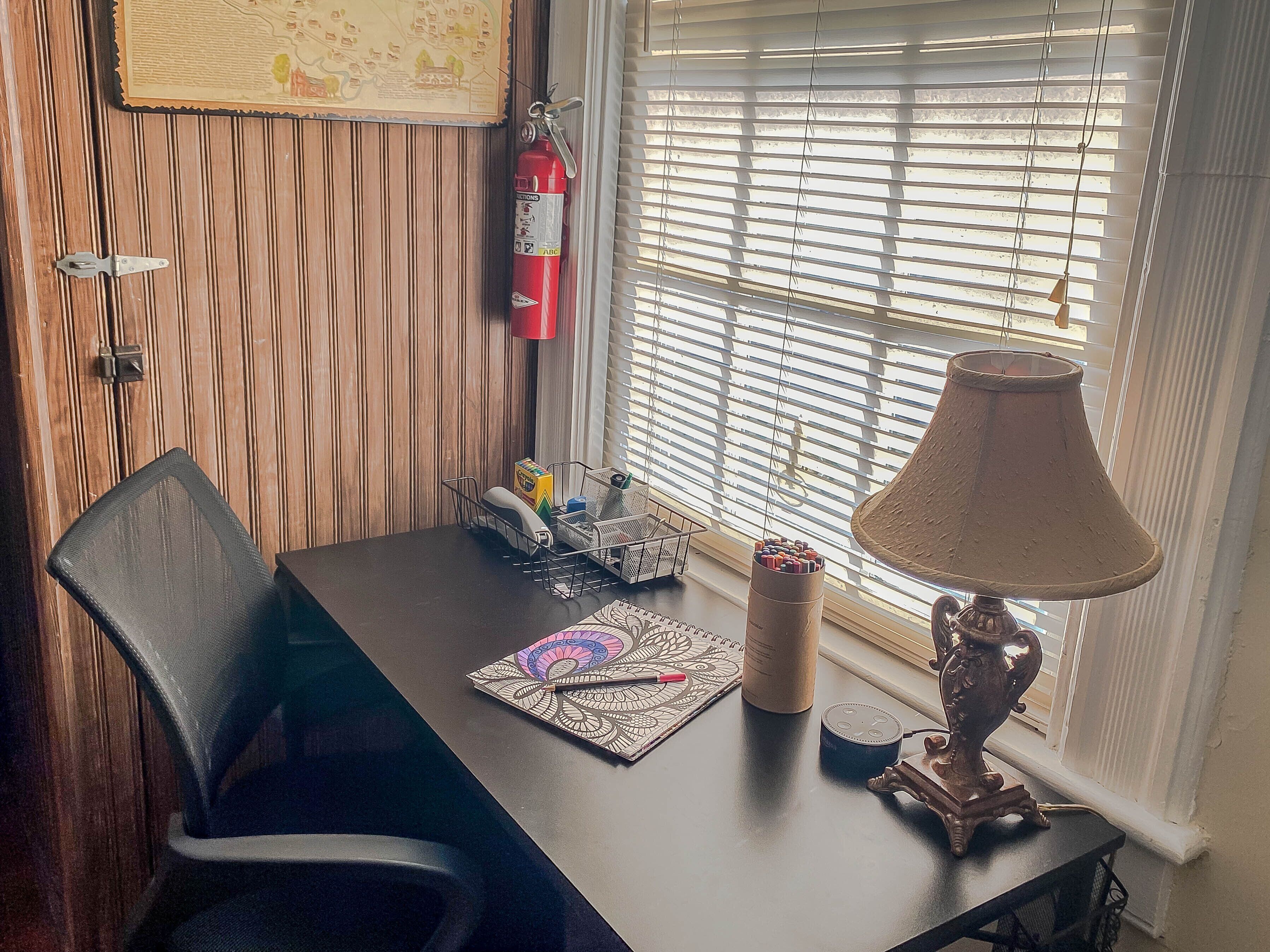 A wooden-paneled room with a desk, chair, lamp, fire extinguisher, and window blinds.