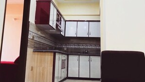 Una cocina compacta con horno integrado, microondas y lavamanos. Hay un mueble de madera con puerta de vidrio y un mueble rojo encima. El techo tiene iluminación empotrada y estructura de vigas de madera.