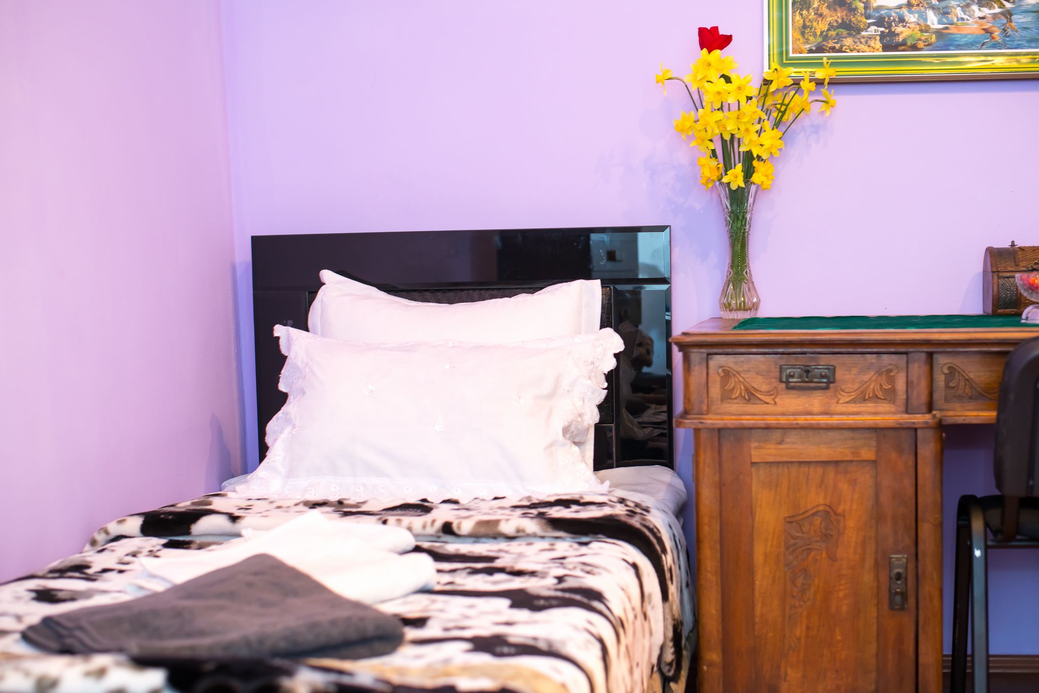 A bed with a patterned bedspread, a bedside table with a vase of flowers, and a wooden nightstand.