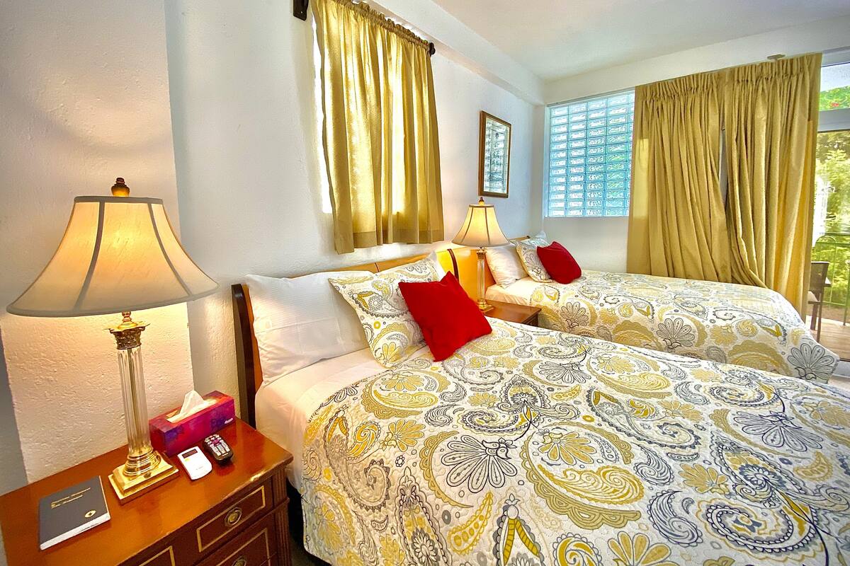 A hotel room with two beds, a nightstand with a lamp, a bedside table with a book and remote control, and a window with curtains.