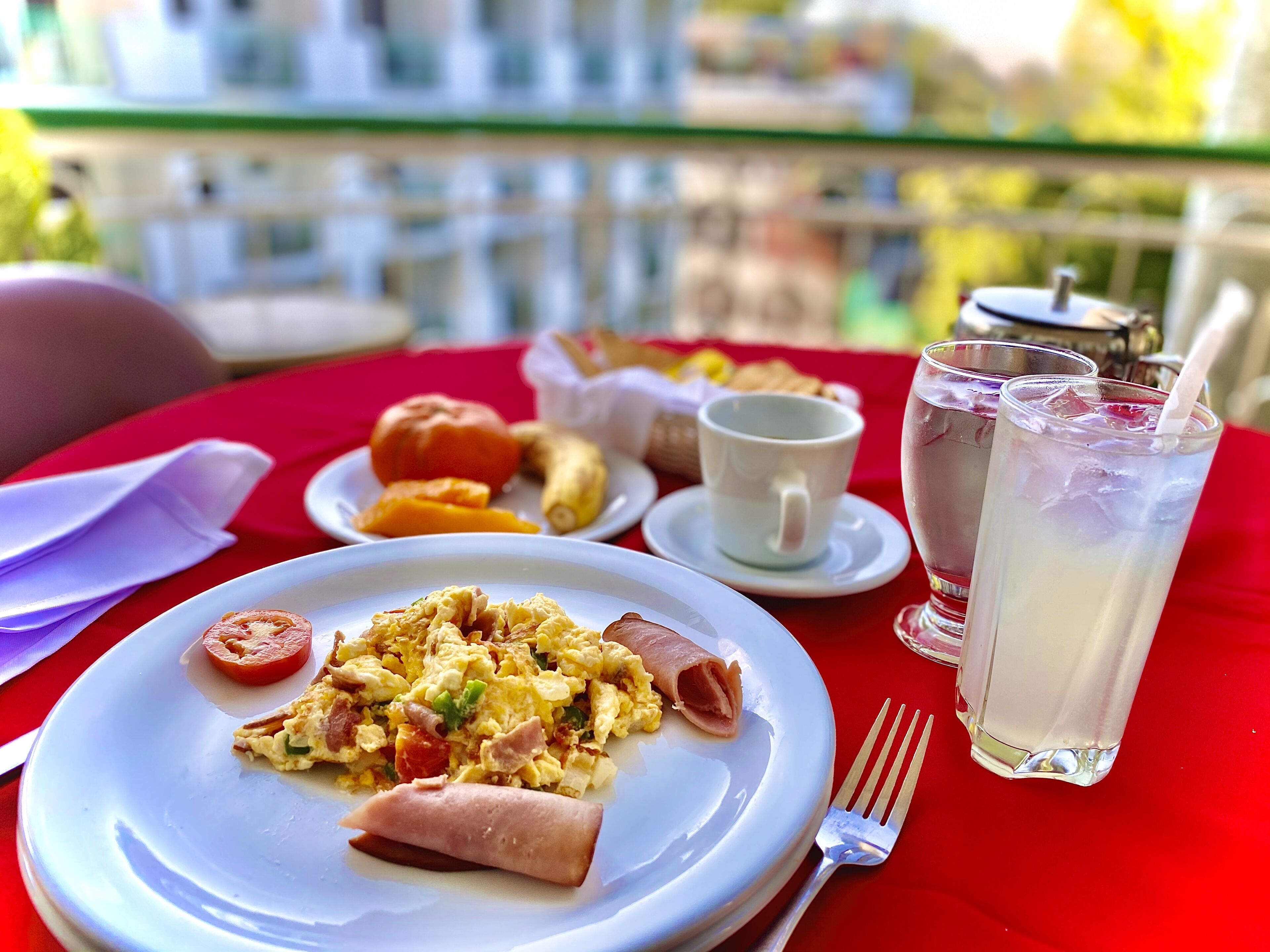 Una mesa de comedor con un plato de huevos revueltos, un panecillo y un tomate. También hay dos vasos de té helado y una taza blanca sobre la mesa.